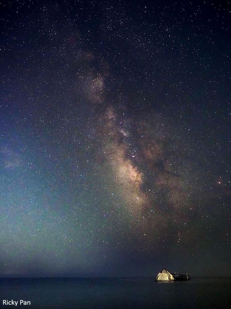 Photo of the milky way with the ocean and a shipwreck appearing above the horizon.